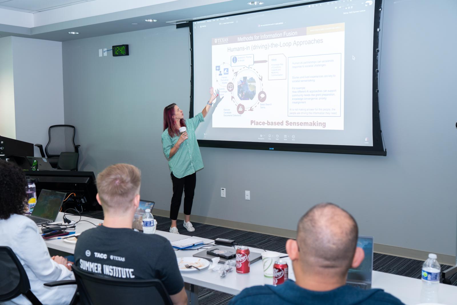 Co-PI Kasey Faust delivers a guest lecture at TACC's Decision Support Office Hands-on Summer Training Institute in May. The slide being projected is titled "Humans-in (driving)-the-Loop Approaches." The backs of three attendees sitting in the front row can be seen in the foreground.